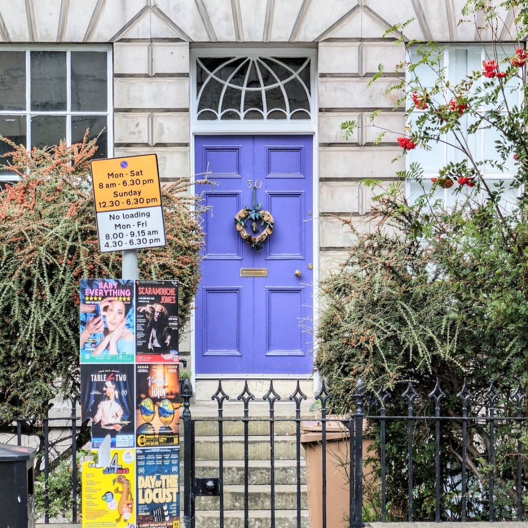 A vibrant purple front door decorated with a wreath on a stone Edinburgh building, framed by lush foliage and a black iron fence covered in colorful Edinburgh Fringe Festival posters.