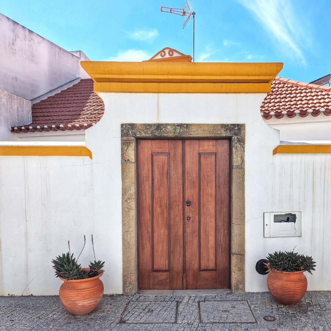 Warm honey-toned wood double door set into a white courtyard wall with mustard-yellow trim and terracotta roof tiles, flanked by potted succulents on Portugal's Alentejo coast.