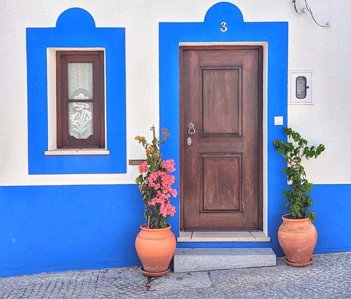 Rich brown wood door numbered 3 in a blue-trimmed white archway, with a matching blue-framed lace-curtained window and pink bougainvillea in terracotta pots on Portugal's Alentejo coast.