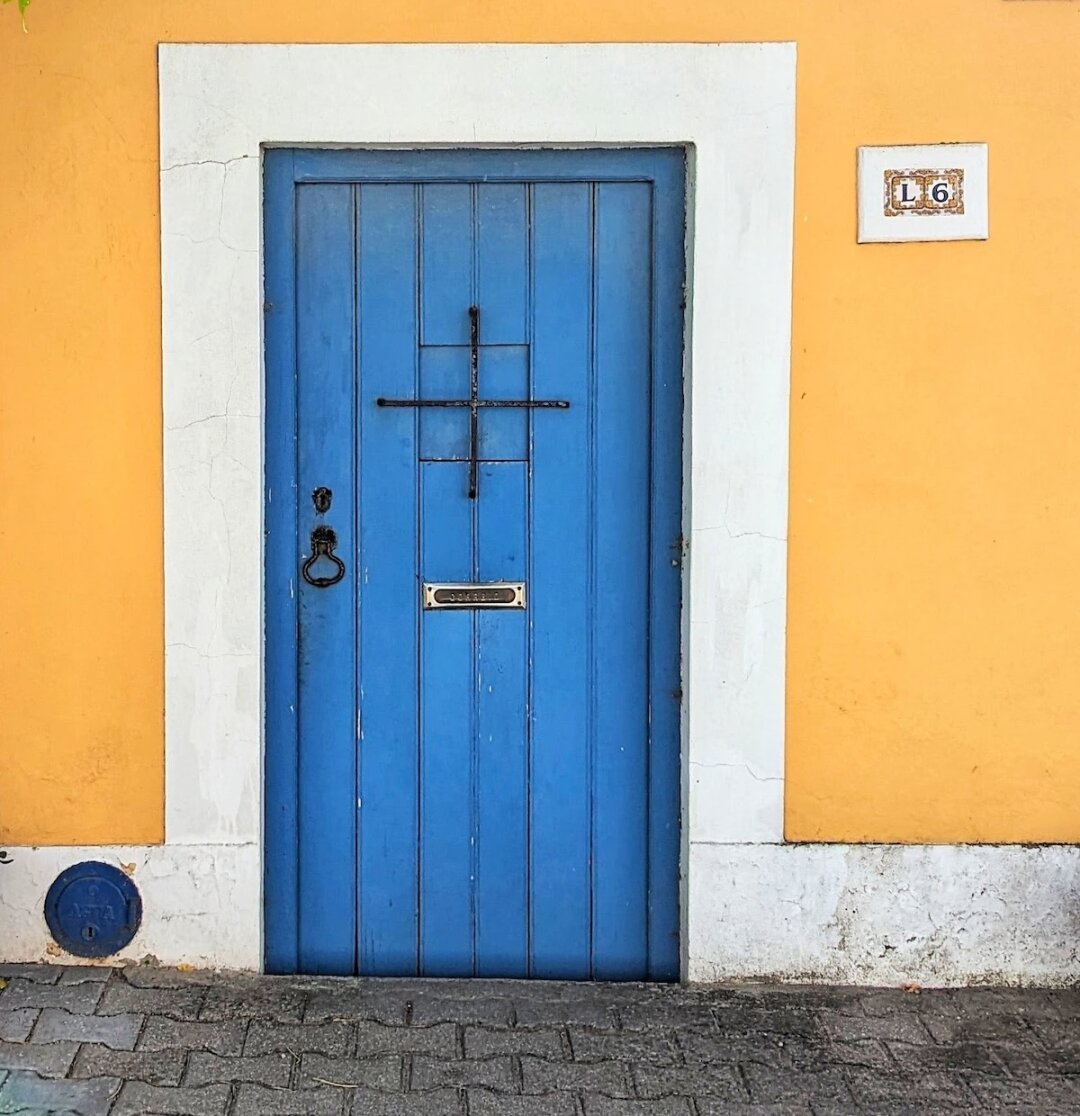 Cornflower-blue plank door with a black cross-shaped iron grille and ring knocker, set in a warm orange wall with a numbered "L6" tile plaque on Portugal's Alentejo coast.