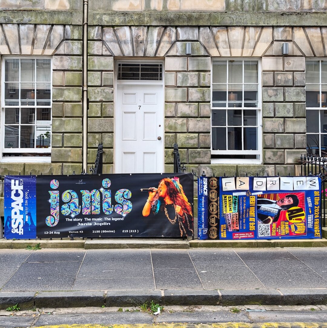 A simple white door and sash windows set into a classic stone townhouse facade, with large Edinburgh Fringe event banners secured across the front railing.