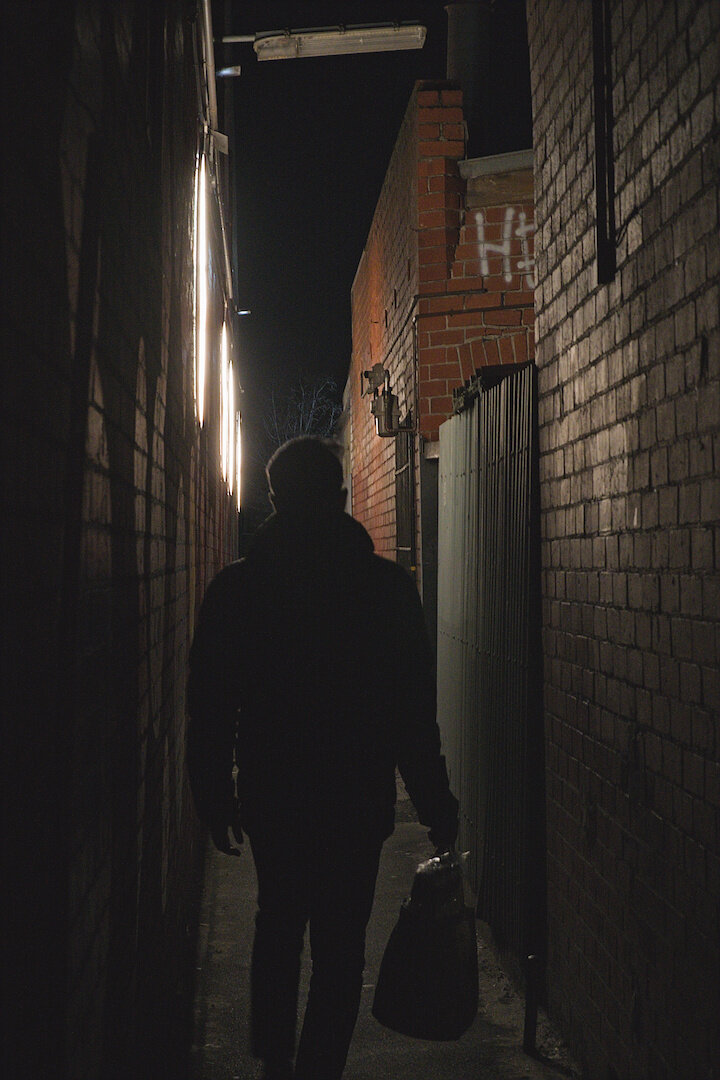 A photograph of a man walking through an alley in the evening. The man is facing away from the camera and is back-lit, only his silhouette is visible. He is carrying a shopping bag in his right hand. The alley is lined with brick walls on both sides, as well as a soft-light-green corrugated metal wall for a section on the right hand side. There is a small amount of graffiti visible high up in white paint spelling the letters "HI" or perhaps "HE" (cut off in the photo). The left wall is lined with bright circular lights which are reflecting on the bricks on the right wall. The sky is dark - it has just turned night and while not pitch black, there is a small amount of blue remaining. A tree with no leaves can be seen in the distance beyond the end of the alley. There are pipes and gas meters in the alley. There is a bar lamp overhead which was off at the time the photo was taken but was flickering at the time. The left wall is painted but it is hard to tell what as it is dark.