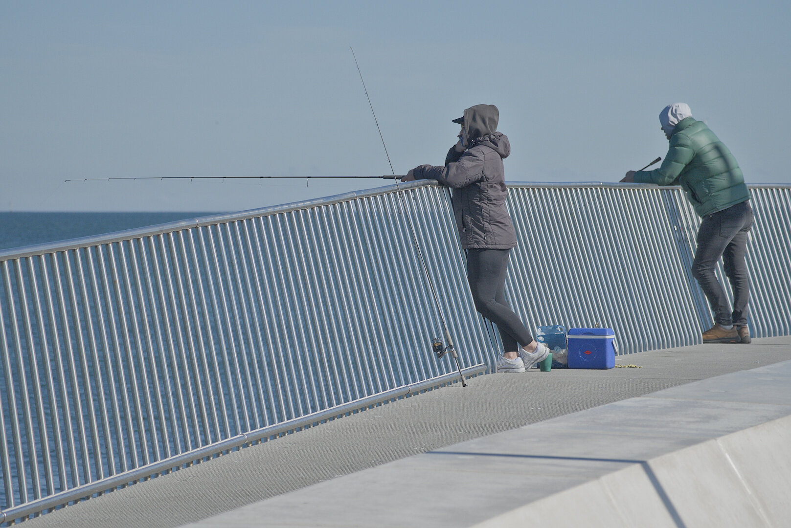A photograph of two men fishing over the railing of a pier. The photograph is taken at a distance. One man is in the centre of the frame and the other is on the far right. They are both wearing warm clothing. The sky is clear and a little hazy. The pier railing is made of metal bars, with a horizontal bar along the top and the bottom and evenly spaced vertical bars between creating a repeating pattern. The ocean can be seen behind the bars and the horizon line can be seen a little above the fence on the left of the image. The lower right of the image is mostly taken up by the footpath of the pier and a concrete structure. It's mid-afternoon in winter, and the scene is lit with low and soft winter sun from the back-right.