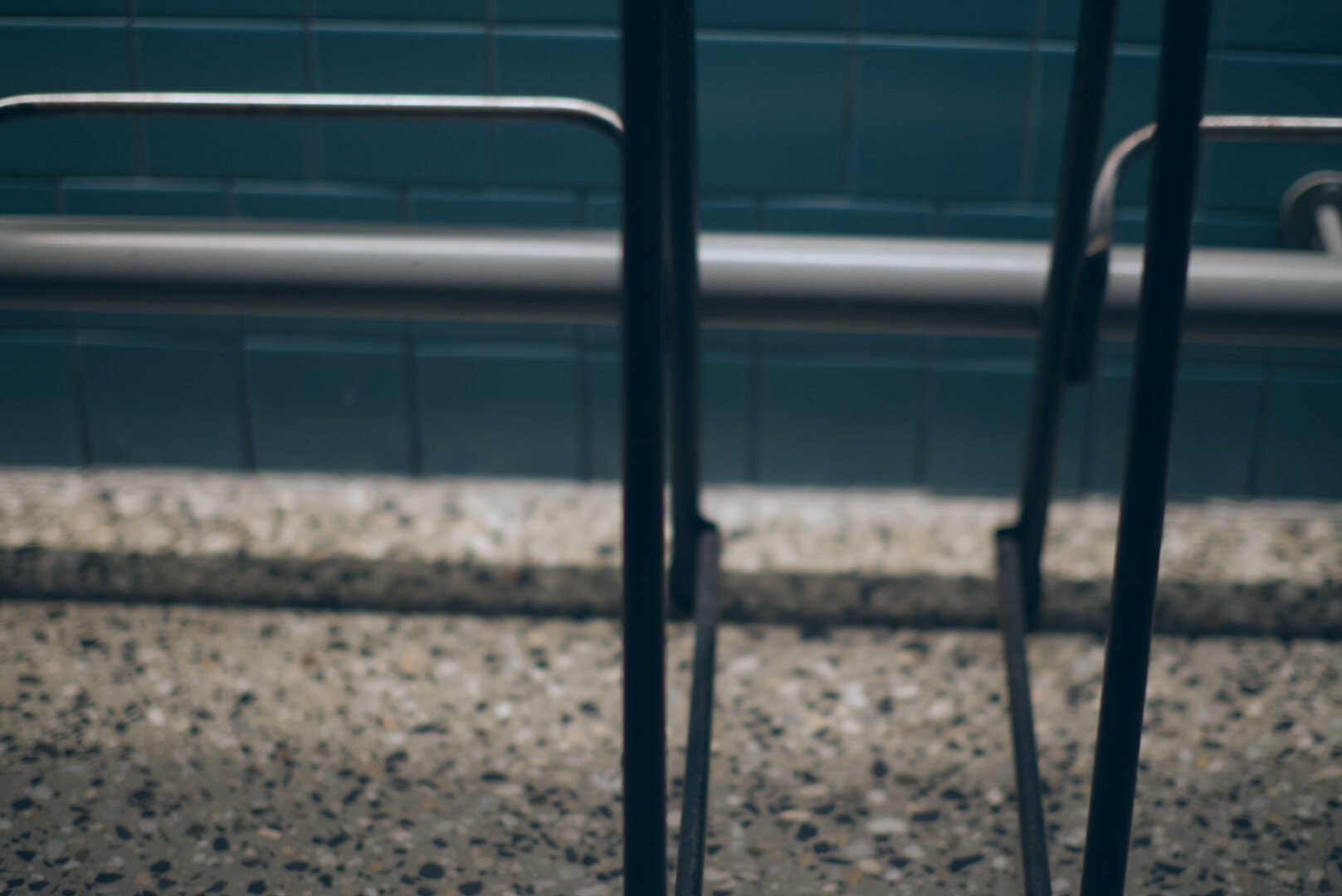 A photograph looking at the bottom half of an aqua-coloured tiled bar wall in a restaurant. There is a metal bar footrest running horizontally. There are the frames of two black metal bar stools partially in view. The floor is polished concrete showing stones of various colours scattered throughout the concrete. The photo has a soft focus and soft lighting is eminating from above casting a shadow on the foot bar down to the ground.