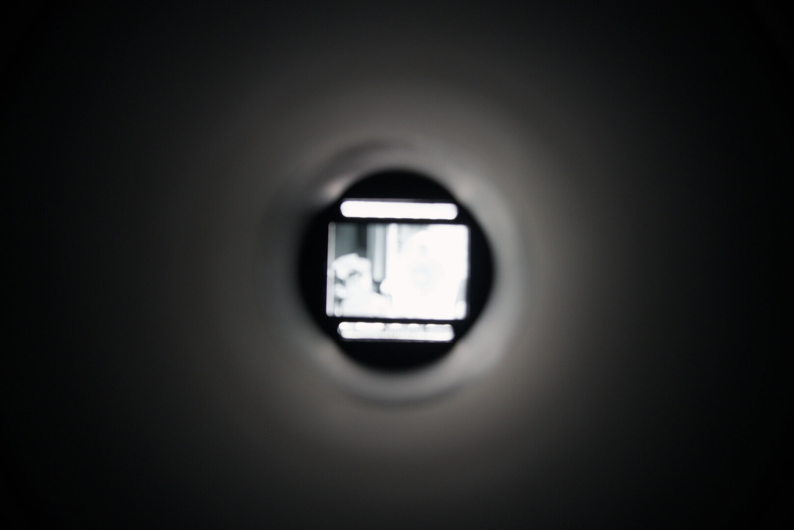 A photograph of a film negative through an extension tube. The tube takes up most of the frame, and is dark around the edges and gradates towards a lighter grey in the middle. In the centre of the tube is the film negative. It is overexposed and out of focus. Above and below the negative are the two bars which usually contain information and barcodes. These are also out of focus, but you can barely make out what appears to be a barcode in the lower bar. The image in the negative is not particularly distinguishable, there are dark details in the upper left corner, some vertical bars in the centre, and a few other patchy areas but nothing particularly noteworthy.