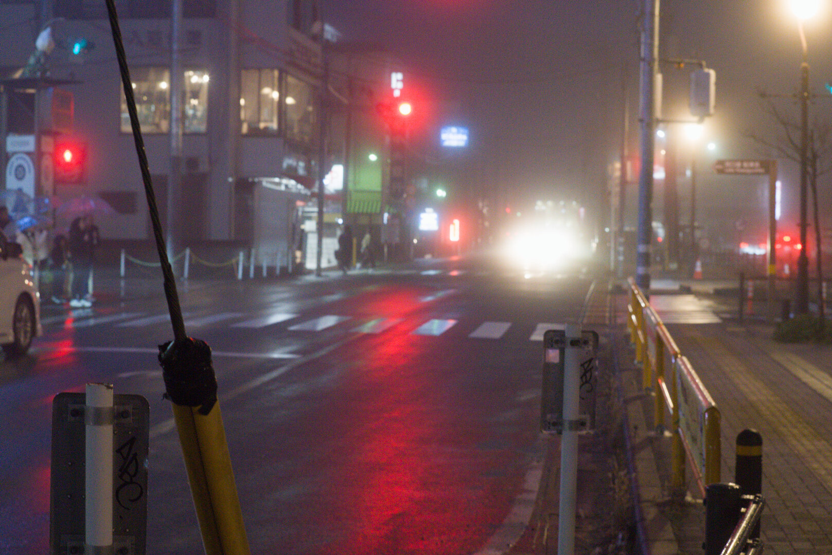 A night photograph of a street in light fog, with a set of traffic lights and a pedestrian crossing centre-frame. There are lights everywhere including the red traffic lights, as well as blooming bright white lights from a bus in the distance. The road is wet and reflective amplifying the light. Around seven pedestrians waiting on the left side of the road ready to cross.