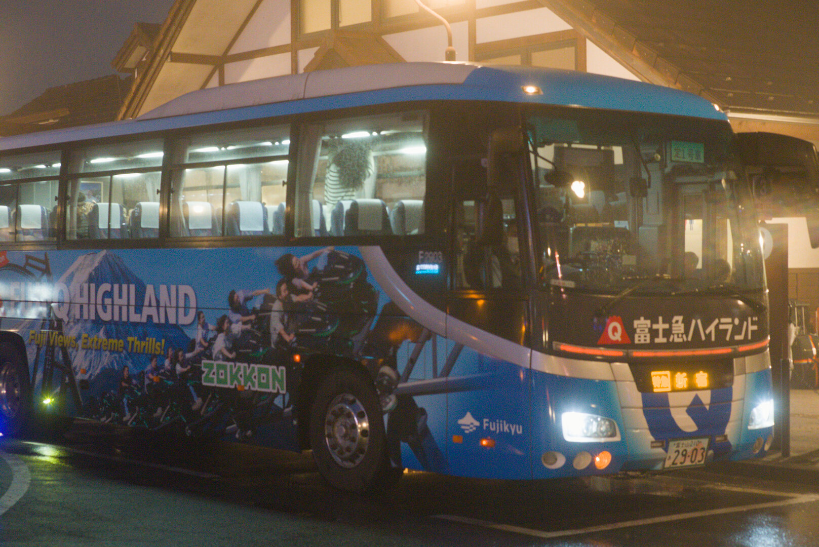 A night photograph of a coach bus in light fog. The bus is wrapped in advertising print for "Fuji Q Highland - Fuji Views, Extreme Thrills" and pictures a roller coaster full of people with Mt Fuji in the background. On the bus there is one person in the middle reaching up to load or unload an item from the overhead storage bins who is wearing a black-and-white striped shirt and has long curly hair, a few more people towards the back of the bus who are hard to distinguish, and the driver. Behind the bus is a building bathed in yellow light. The building is mostly white with a brown wooden frame so it readily shows the light reflecting on it.