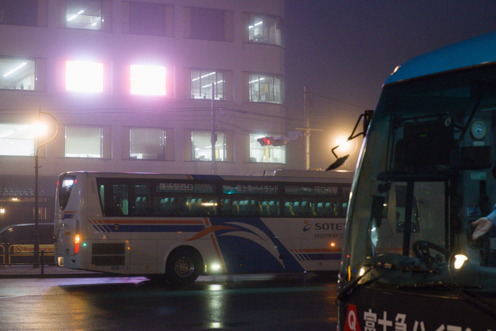A night photograph of a street in light fog. There is a multi-story building in the background which appears to be an office or apartment building. Most windows are illuminated and two square windows are glowing particularly brightly with a purple hue - you cannot see anything else in them. At the road level there is a coach bus on the opposite side of the road, and in the foreground on the right there is the front of another coach bus. The arm of what I presume is the driver can be seen gesturing, perhaps inviting a passenger to board or disembark. There are street lights on the opposite side of the road which glow like small suns and are suspended on thin poles that hook around the outside of the globe.