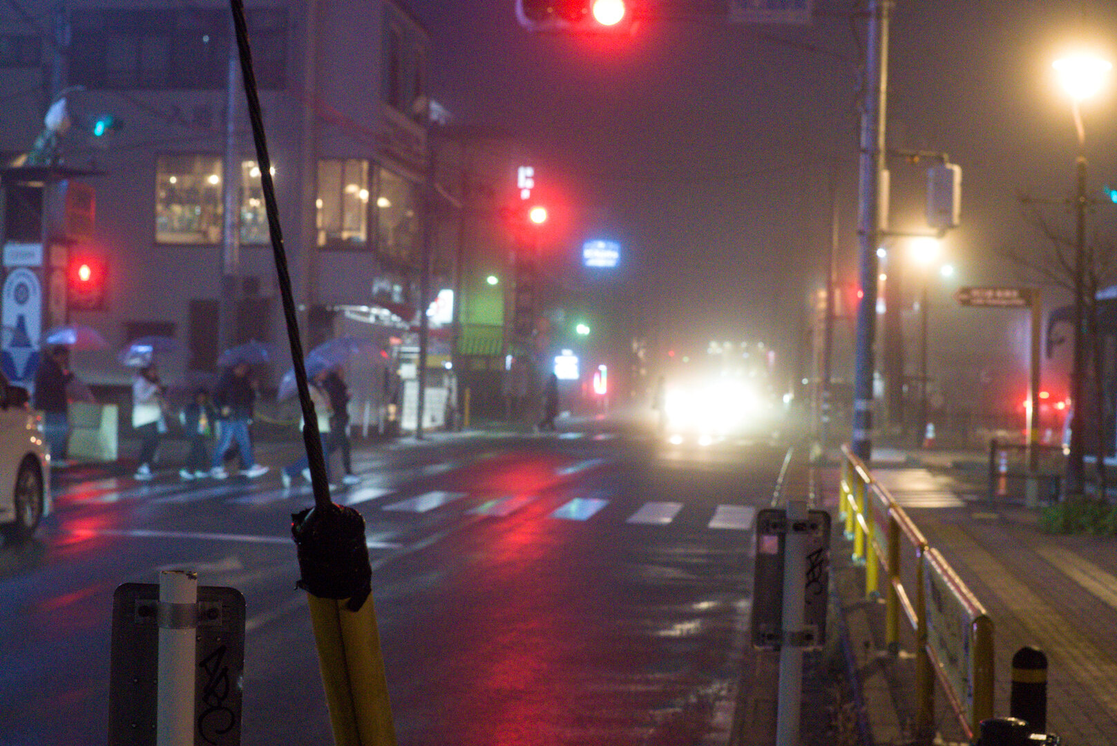 A night photograph of a street in light fog, with a set of traffic lights and a pedestrian crossing centre-frame. There are lights everywhere including the red traffic lights, as well as blooming bright white lights from a bus in the distance. The road is wet and reflective amplifying the light. Around seven pedestrians are crossing from left to right. Most pedestrians are carrying transparent white PVC umbrellas.