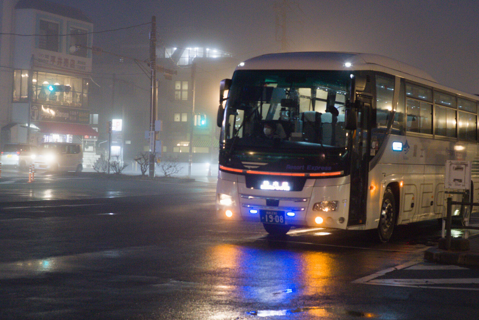 A night photograph of a street in light fog. To the right of the photo is a coach bus facing at a 45º angle. The bus has various lights on the lower front that reflect off the surface of the road, creating yellow and blue gradients. The coach features other lights and signage including a digital destination sign, a horizontal strip light, headlights, side lights, internal lighting, and some bright lights at the top corners. In the background is a small parking lot, some buildings at a medium distance, some power poles, a parking sign, and a pedestrian crossing.
