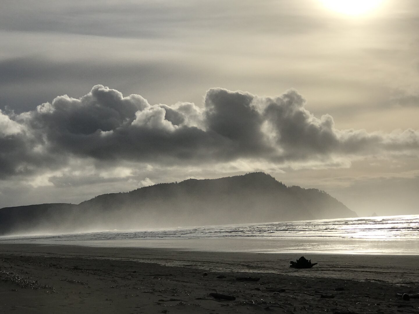 Large clouds above Clark's Mountain and Tillamook Head on background. Sparkling ocean waters in mid ground and strand (beach) on foreground. Entire photo in more or less gray tones.