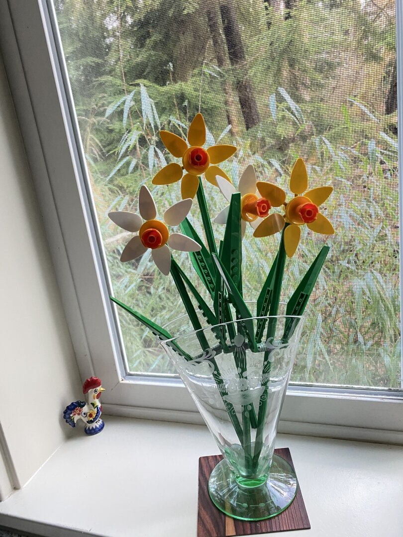 Four Lego daffodils of yellow and orange blossoms, green stems and leaves in glass vase sitting on window sill. Small mostly white "Galo de Barcelos" from Portugal to the left of vase. Trees in background outside of window.