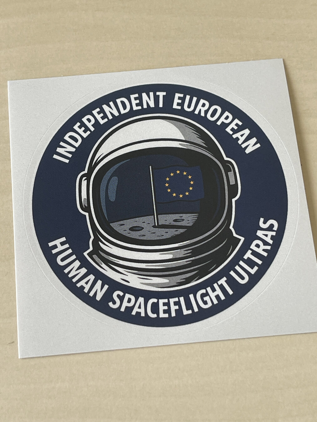 Sticker showing a apace suit helmet reflecting the EU flag on the moon and the caption „Independent European human space flight ultras“