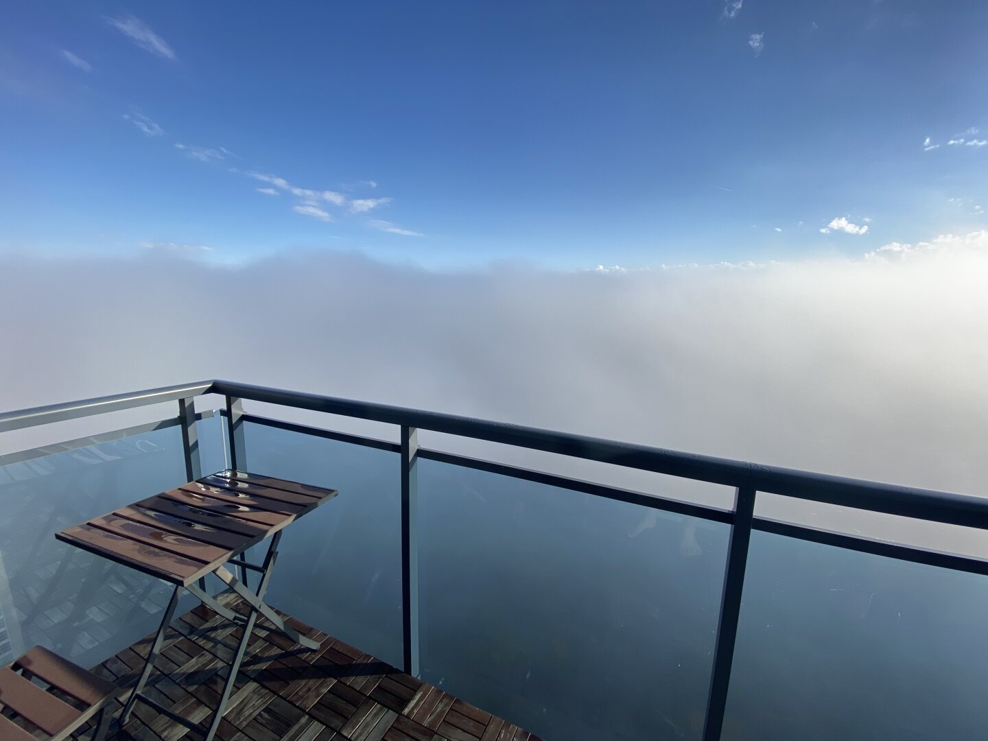 A somewhat surreal view of a corner of a balcony with a table and chair underneath a blue sky. It looks surreal because the balcony is on top of the cloud cover -  like the view from an airplane window.