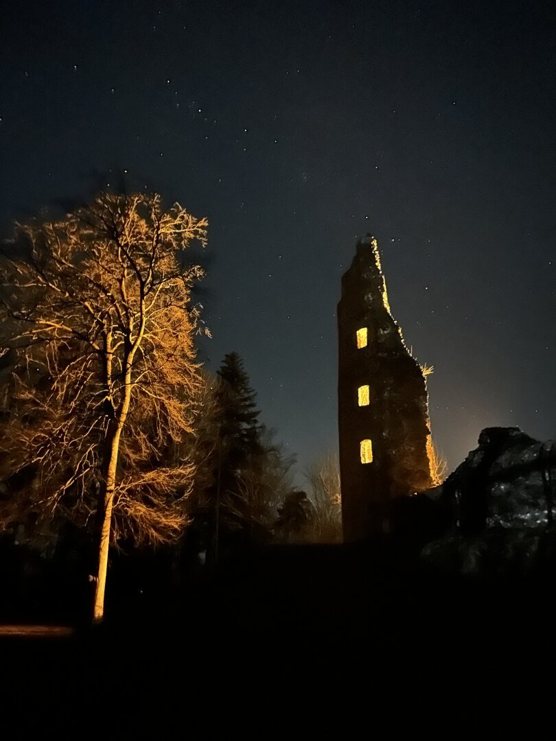 A dramatic night time shot. Beneath a night sky with stars, on the left a tree and on the right a ruined half tower. Inside the tower is a light source shining through three window openings, illuminating the tree on the left. Dramatic effect between the indirect lighting, the silhouette of the tower and the dark sky