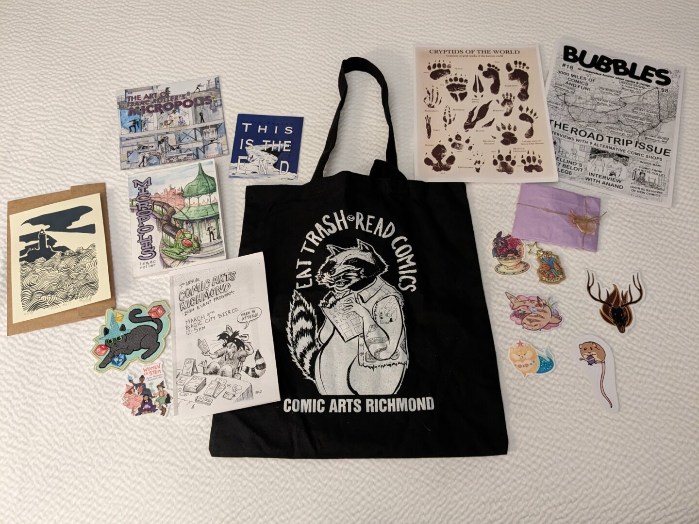 Pile of comics and stickers, center is a black bag with a raccoon in a denim vest reading comics surrounded by the words "Eat Trash Read Comics, Comic Arts Richmond"