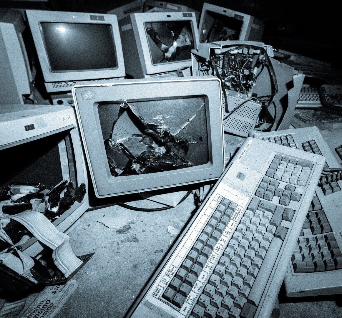 This image depicts a chaotic scene of broken and discarded vintage computer equipment. The foreground shows damaged CRT monitors with shattered screens, tangled wires, and cracked components. Keyboards from older systems, such as IBM or Dell models, are strewn across the scene, some with visible signs of wear and grime. The environment appears abandoned and dusty, suggesting it might be an old technology scrapyard or a deliberate setup for a grungy aesthetic. The bluish tint adds a cold, dystopian mood to the image.