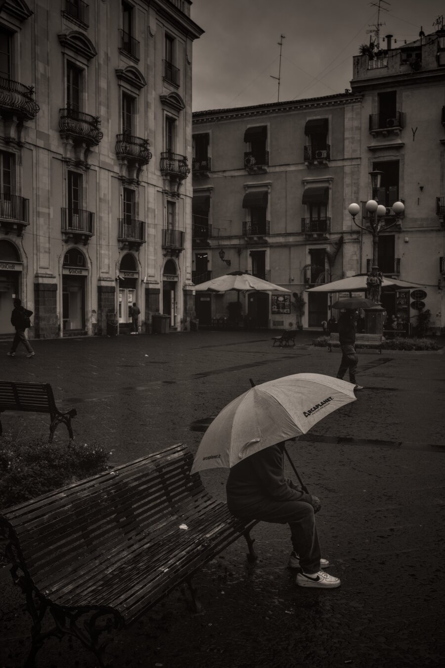A moody, sepia-toned street photo captures a quiet, rainy day in an Italian square. A person sits alone on a bench under an umbrella, while others pass by in the background. Wet pavement, old buildings, and subdued lighting give the scene a reflective, cinematic atmosphere.
