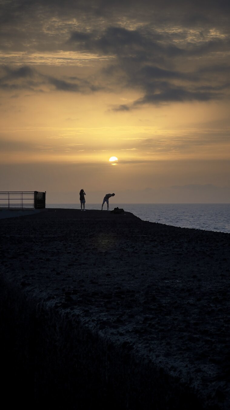 The image depicts a peaceful sunset by the sea, with two silhouetted figures on a rocky promontory. The golden sun, partially obscured by clouds, casts warm hues across the sky, while the calm sea reflects the light. A metal railing borders the platform, adding structure to the serene scene.