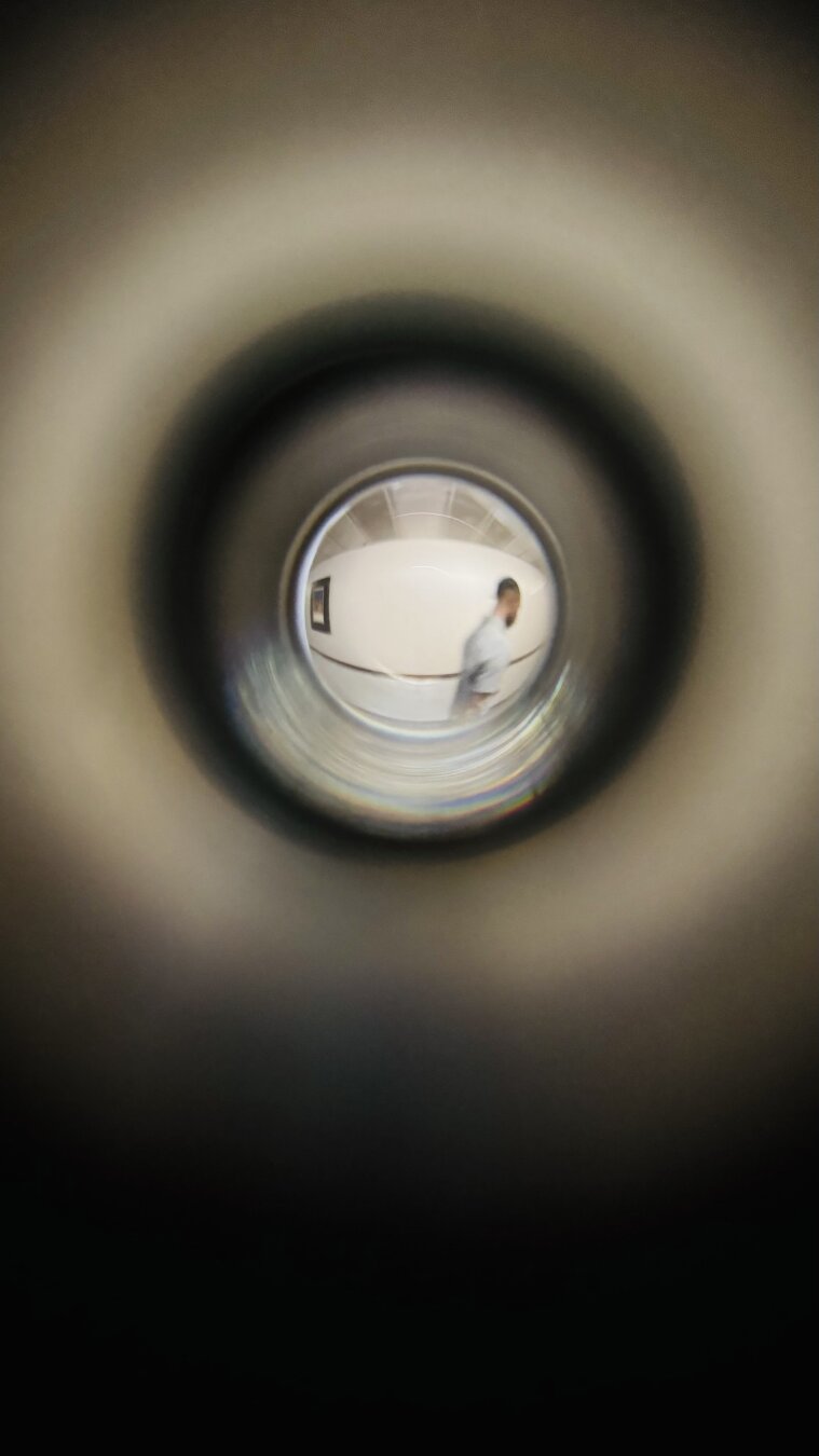 This image appears to be taken through a door peephole (fisheye lens effect), creating a circular and distorted view of the scene outside the door. In the center of the frame, a person is walking through a hallway. The individual is slightly blurred, likely due to motion, and is dressed in light-colored clothing.

The hallway is relatively plain, with light-colored walls, a dark chair rail, and framed artwork hanging on the wall. The image has a vignette effect around the edges, characteristic of the narrow, curved field of view seen through a peephole. The overall perspective gives the impression of someone discreetly observing from inside a room.