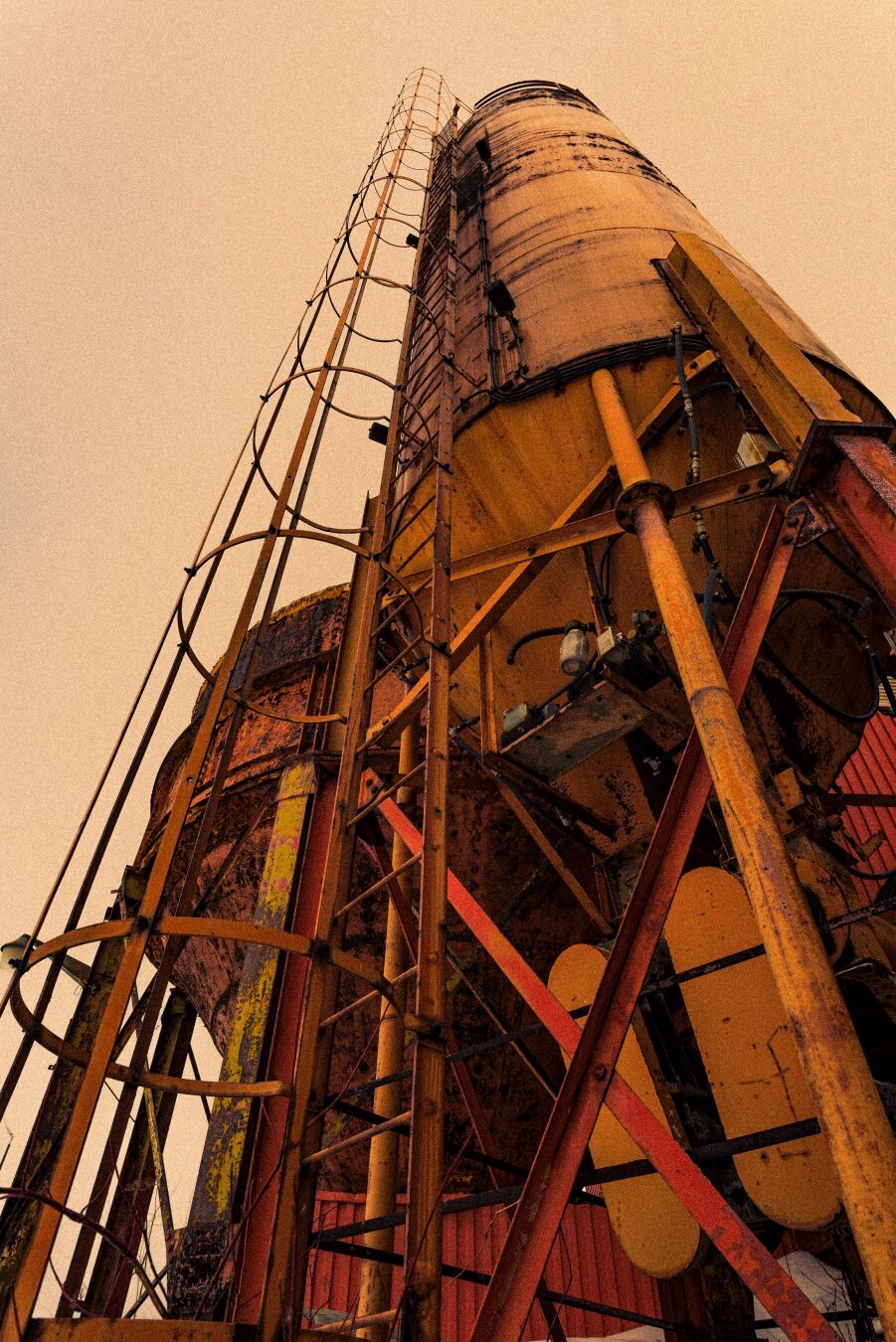 This image captures a striking industrial scene, focusing on a rusted, weathered silo structure shot from a low angle. The ladder and surrounding framework, composed of yellow and orange steel, add a sense of geometry and depth. The warm, gritty tones give the photograph a vintage, almost post-apocalyptic aesthetic, accentuating the corrosion and wear on the metal surfaces. The background features parts of a red structure, enhancing the overall industrial theme. This composition effectively conveys a sense of age, decay, and rugged beauty.