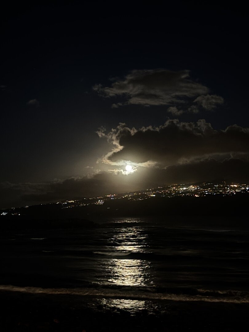 A nighttime seascape featuring a bright moon shining through clouds, reflecting on the water. Distant city lights are visible along the coastline.// Un paisaje marino nocturno con una luna brillante que brilla a través de las nubes, reflejándose en el agua. Las luces lejanas de la ciudad son visibles a lo largo de la costa.