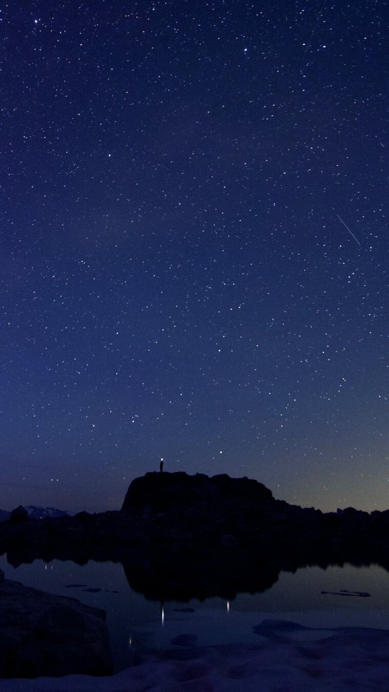 A person standing on a small hill made if rocks at night, with a small lake in front of it, reflecting the stars above