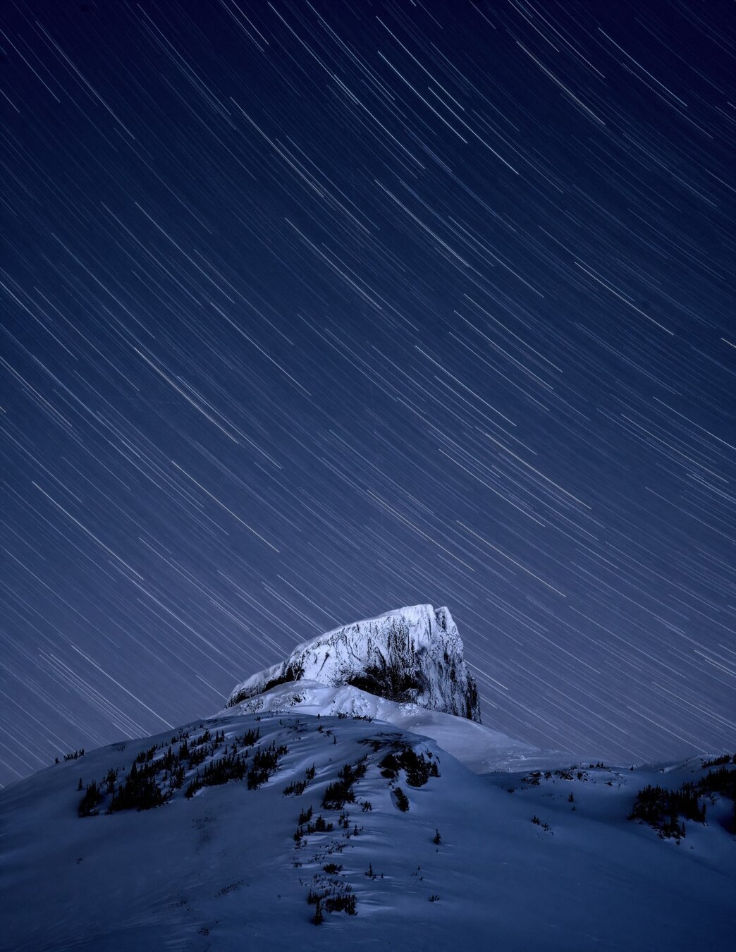 A prominent mountain feature at night capped in snow, a long exposure with star trails behind it.