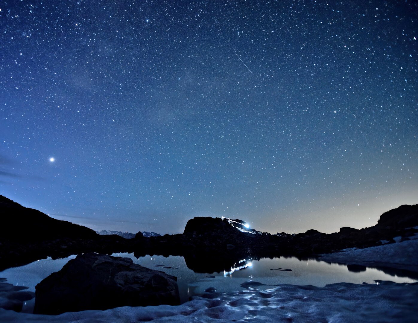 A mountain lake reflecting star light and the light of a headlamp as someone walks along the edge during a long exposure