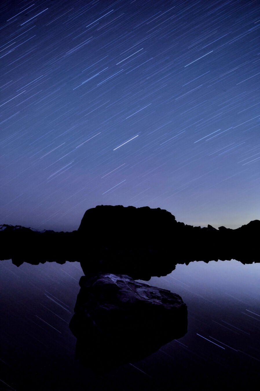 A mountain lake reflecting star trails from a long exposure, with star trails up above