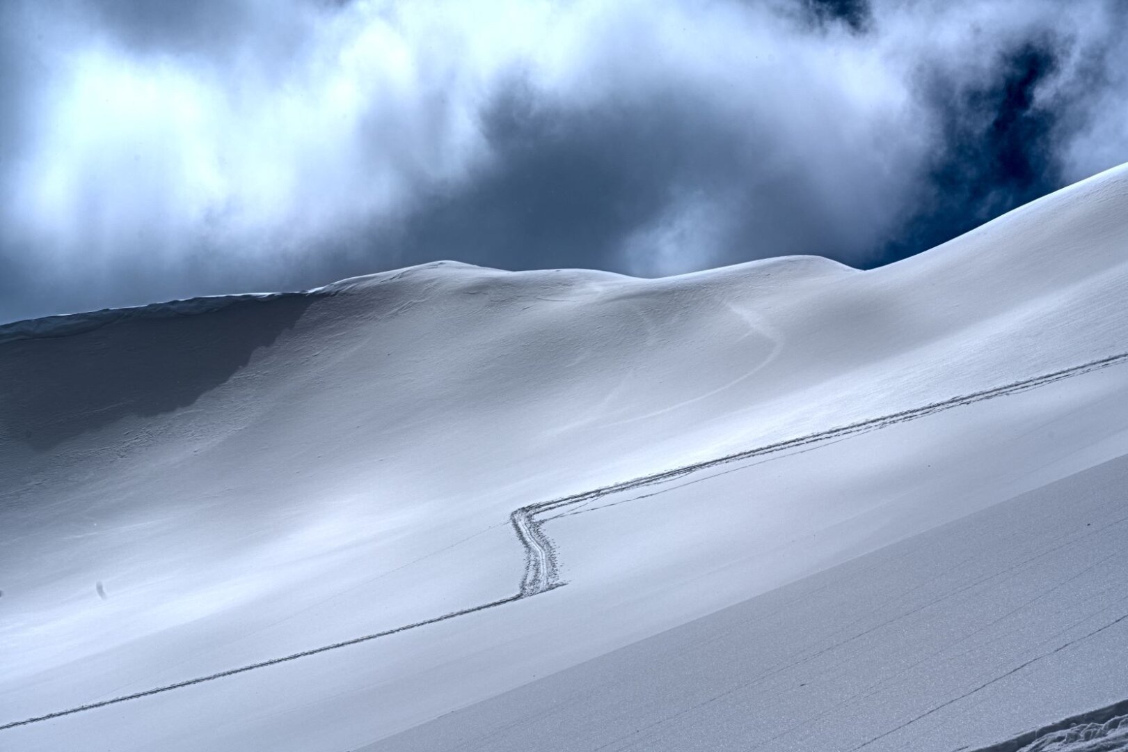 a snow covered mountain scene with a mountain ridge covered in snow and fluffy clouds immediately above it, a single ski skin track cuts the snow on it's way up