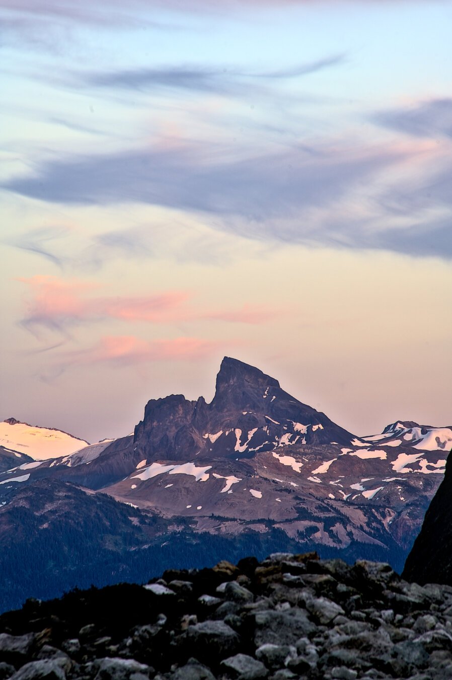 A sharp angled mountain feature, a remnant of ancient volcanic activity, soft clouds above lit as the sun goes down, patches of snow here and there, trees at the base