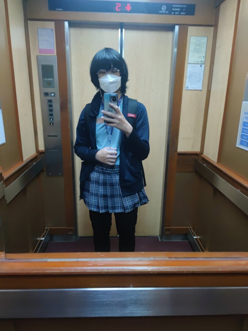 Me holding a phone taking a photo inside of an elevator, I'm wearing a plaided skirt, white shirt and black jacket