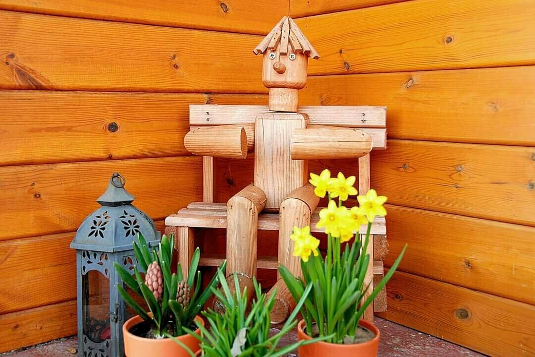 Decorative wooden figure sitting on a bench. Surrounded by potted plants and a lantern.