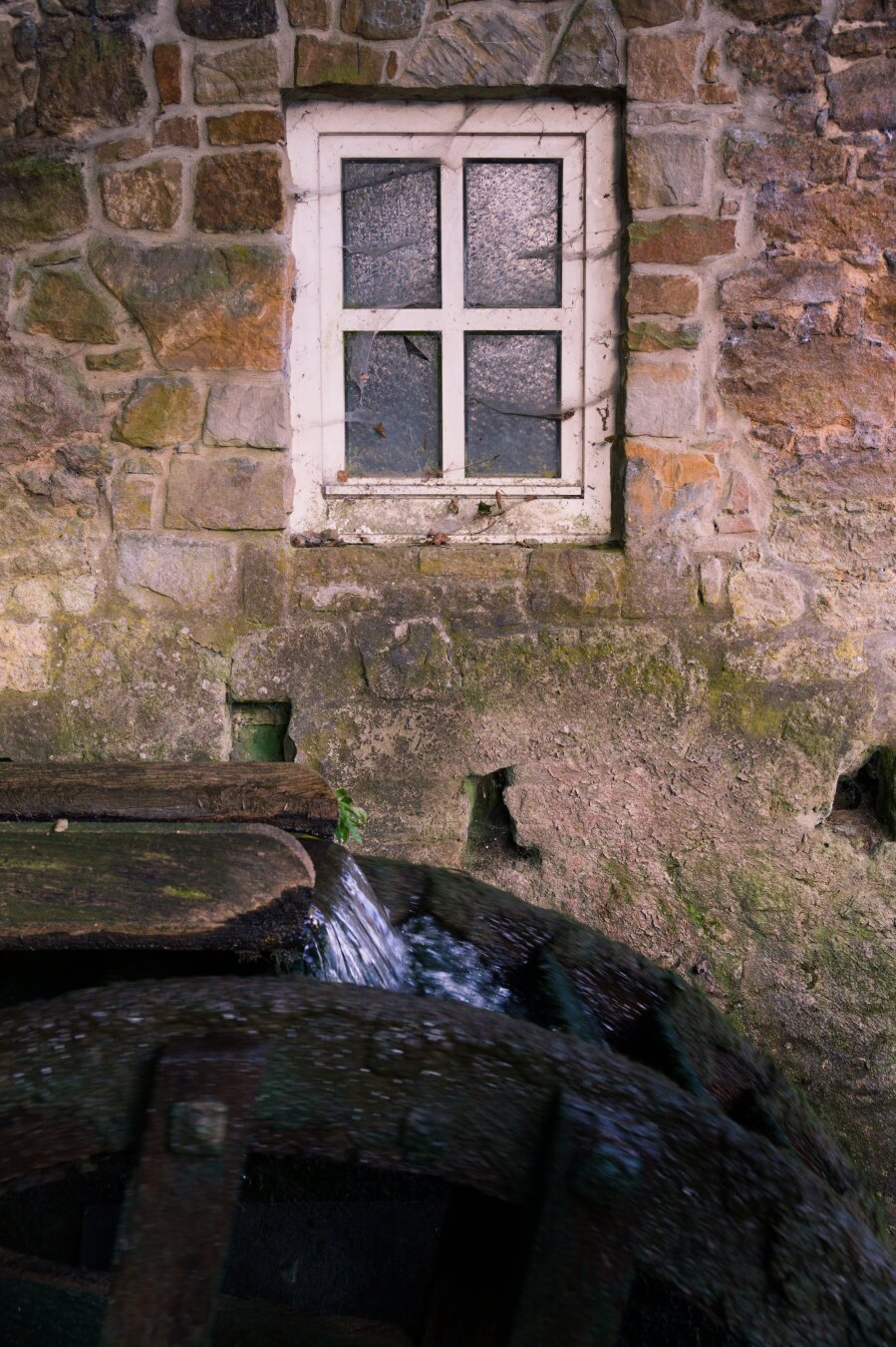 A vertical, medium shot of a rustic stone building featuring an old wooden water wheel. The upper half of the image shows a wall constructed from irregular, earth-toned stones in shades of tan, gray, and brown. In the center of the wall is a small, white-framed window with four textured glass panes. Below the wall, a large, weathered wooden water wheel sits in a shaded area. Water pours from a wooden chute, splashing into the spokes of the wheel. The wheel is dark, wet, and shows significant wear and moss. The lighting is soft, highlighting the rough textures of the stone wall and the aged timber of the water wheel, evoking a historical, industrial atmosphere.