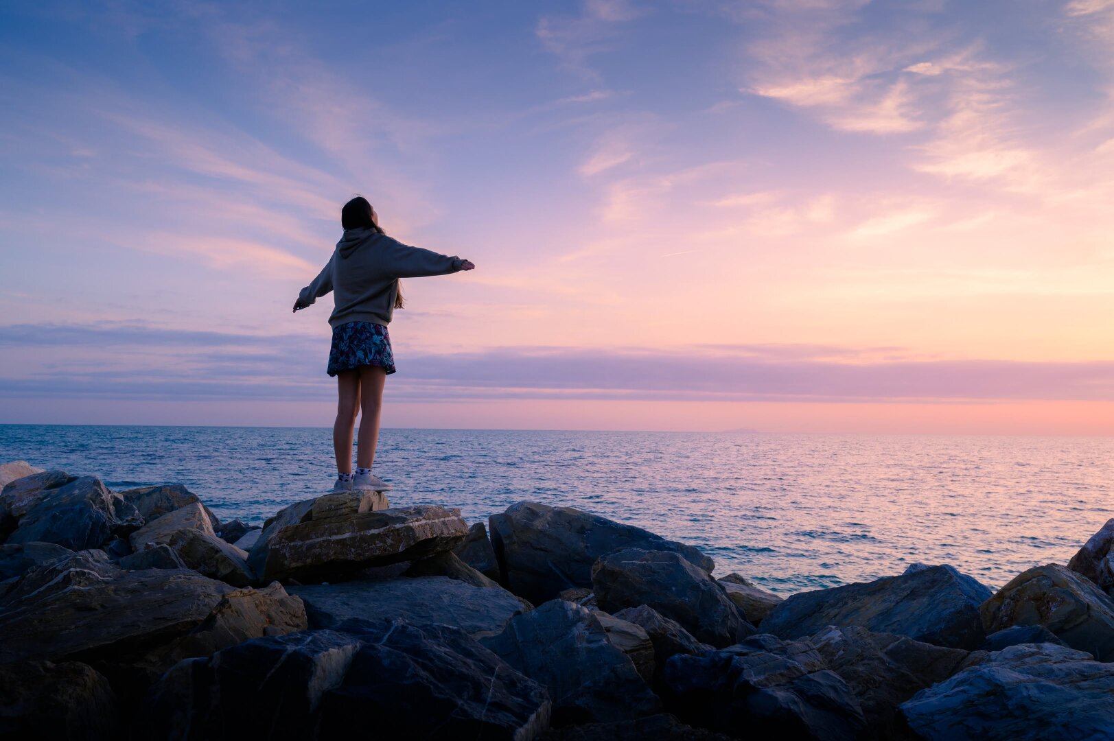 A young person stands on a large, flat rock atop a jagged shoreline of dark gray boulders, facing away from the camera toward the vast ocean. They have their arms outstretched to the sides in a gesture of freedom or embrace. The individual is wearing a gray oversized hoodie and blue floral shorts. The scene is set at sunset, with the sky filled with wispy clouds painted in vibrant shades of deep purple, soft pink, and glowing orange. These colors reflect across the surface of the calm water, creating a peaceful and awe-inspiring atmosphere.