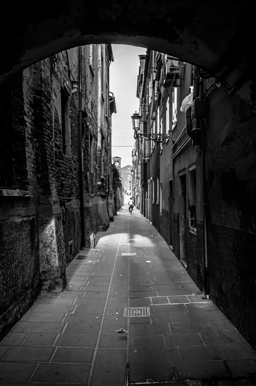 A high-contrast, black and white photograph of a narrow, old-fashioned alleyway viewed through a dark, heavy stone archway. The archway creates a dramatic frame, casting the immediate foreground into deep shadow. The ground is paved with rectangular stones that reflect a bright pool of light in the center of the path. Tall, weathered buildings with classic European-style architecture line both sides of the alley. On the right side, vintage-style street lamps are attached to the walls. In the far distance, a lone, small figure walks away toward a bright opening at the end of the path. The image has a cinematic, moody, and mysterious atmosphere.