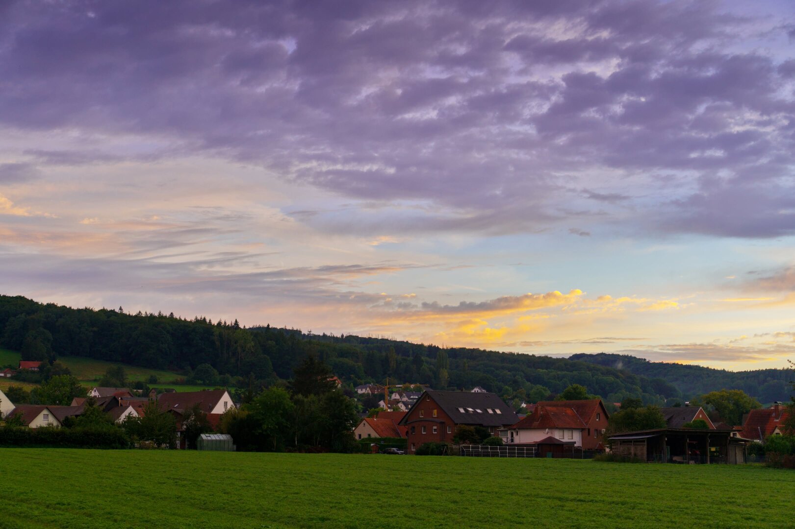 A wide landscape shot of a rural village at twilight. In the foreground, a vibrant green grassy field leads to a row of houses with diverse architecture, including some with distinctive red-tiled roofs, nestled among green trees. Behind the village, a dense, dark green forest rises up a gentle hill. The expansive sky is filled with thick, textured clouds in shades of deep purple and grey, illuminated by golden and orange hues from the setting sun near the horizon.