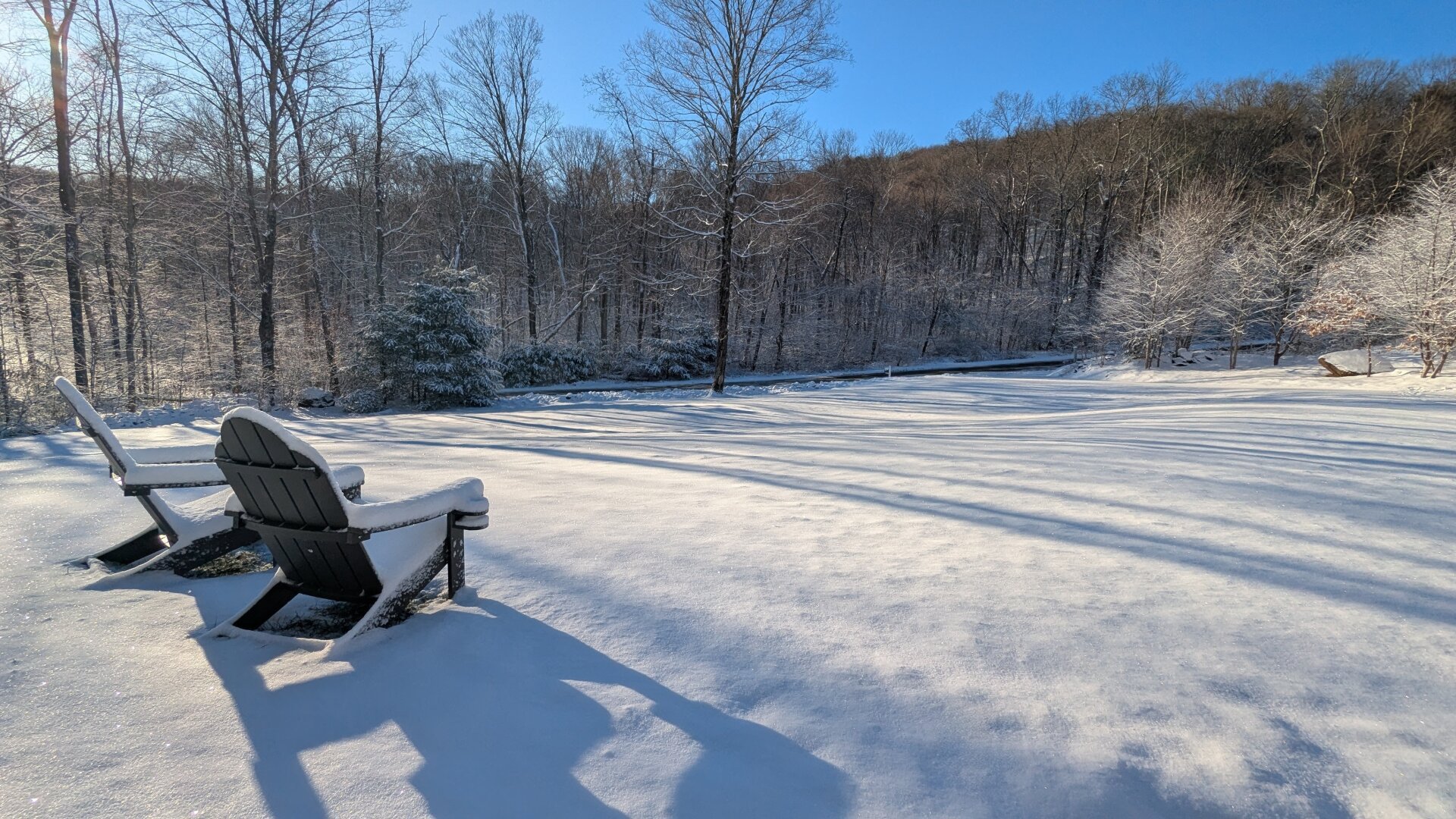 Two snow covered Adirondack chairs on a front lawn which is also covered by snow. The sun is shining from the left side of the photo.