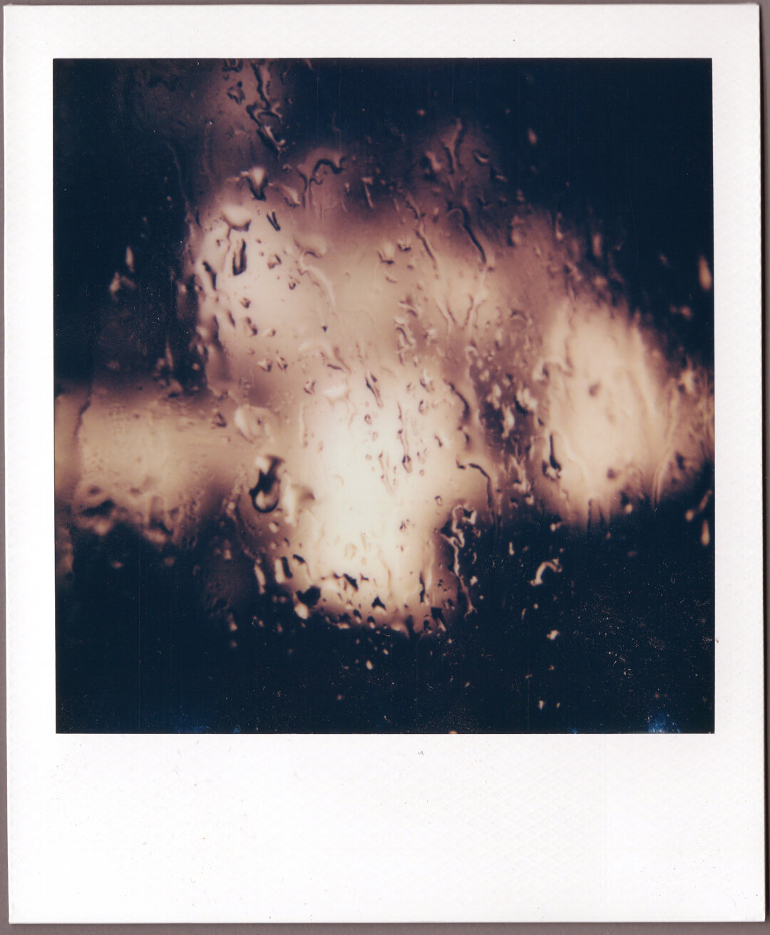 Polaroid, closeup of raindrops on a window, illuminated by indistinct blurry blobs of light in the distance.