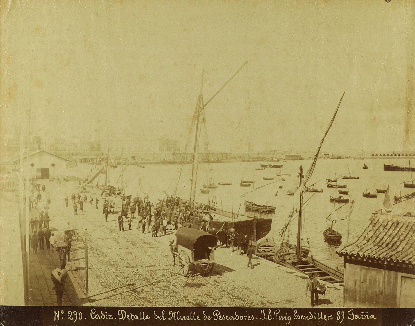 Fotografía amarillenta donde los lugares lejanos aparecen difuminados pero los cercanos están muy bien definidos. Muestra un muelle del puerto con mucha gente, especialmente frente a un barco grande de un mástil.