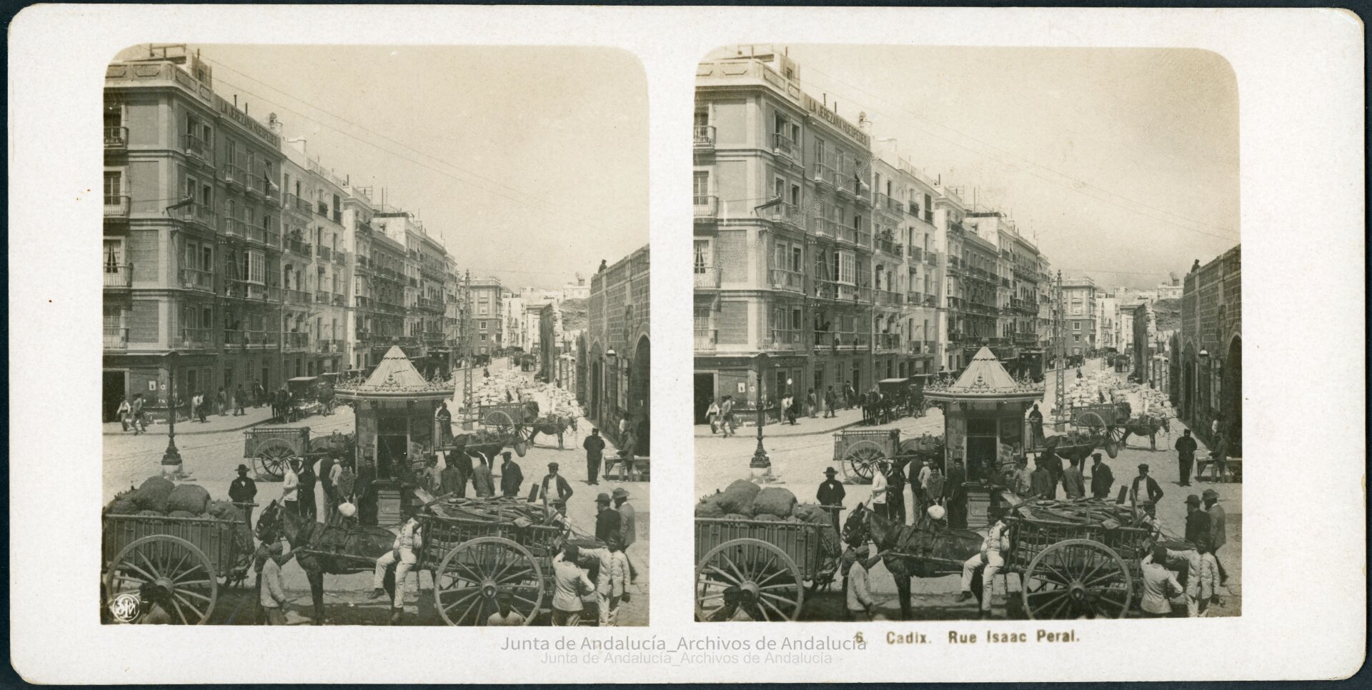 Calle con un quiosco en el centro y mucha gente posando junto a él. Por delante entran carros tirados con burros. Por detrás, salen. En la calle, junto a la acera, están los carruajes y por detrás se acerca el tranvía. A su derecha, la muralla está siendo demolida. A su izquierda, el primer edificio dice en su azotea "La Jerezana Huéspedes", mientras en su esquina inferior hay una tienda de comestibles, cervezas y vinos.