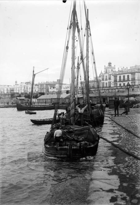 Barco de un mástil amarrado en el puerto. Dos tripulantes miran de frente, sobre la carga, a la cámara. Otro está de espalda en la popa.