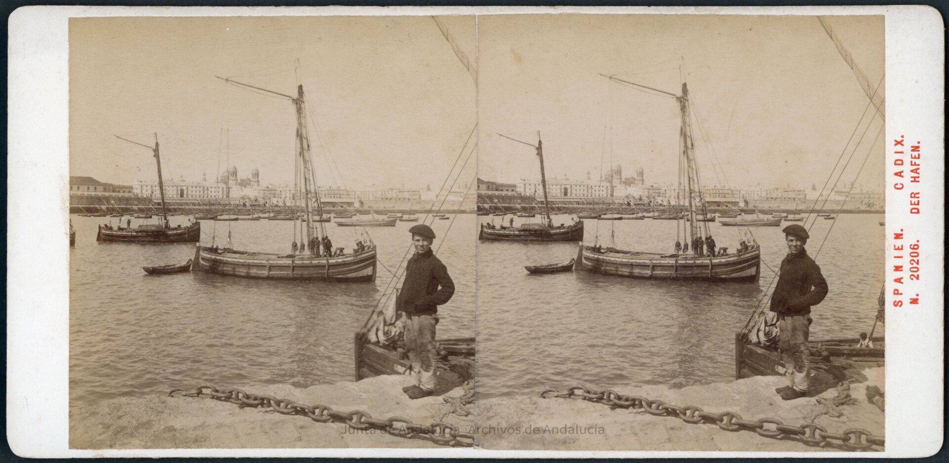 Joven mirando a la cámara desde el borde del muelle. En el puerto hay dos veleros a distancia media, con muchas embarcaciones acumuladas en el fondo, donde se ve la ciudad.