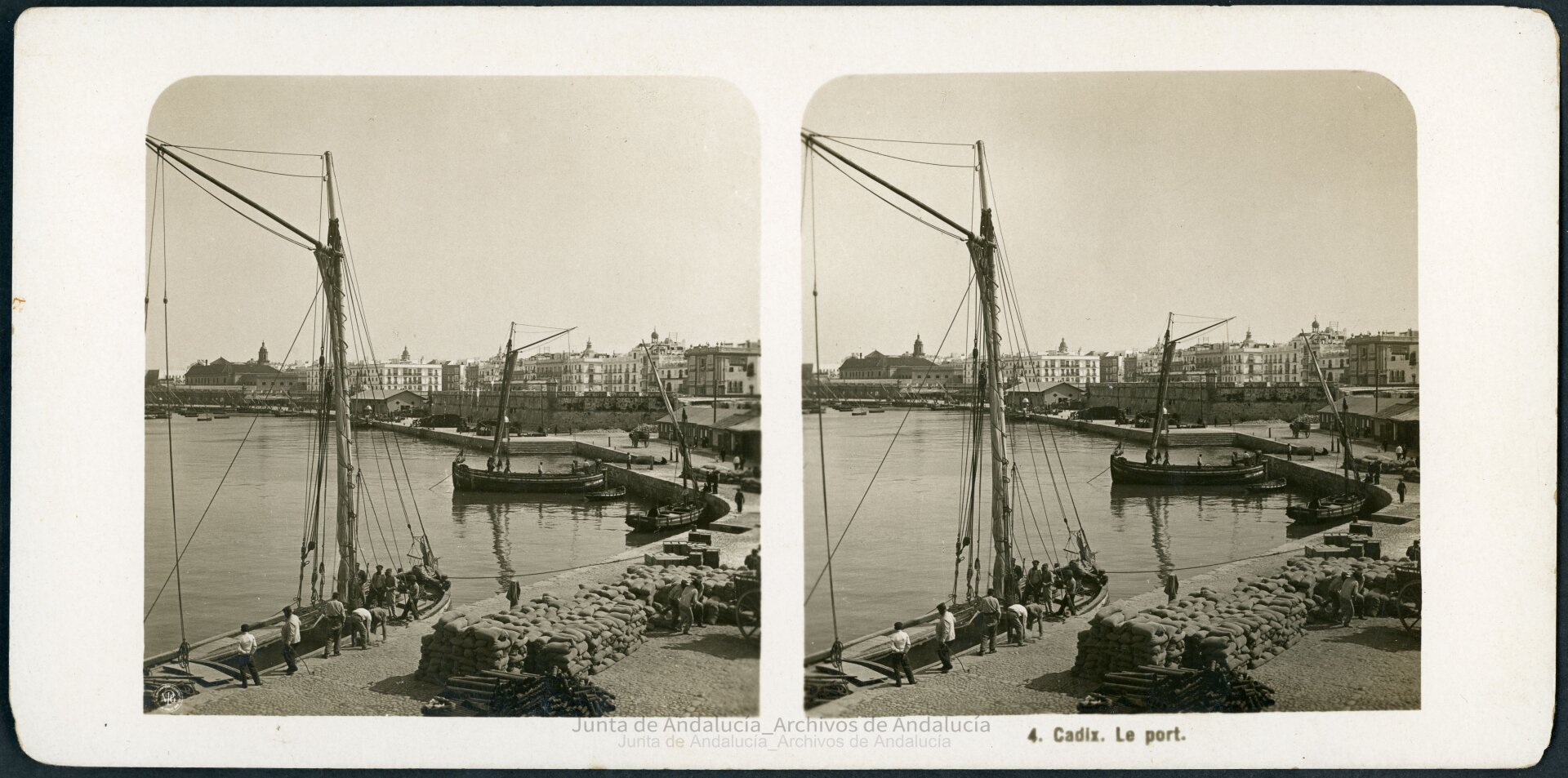 Puerto rodeado por una muralla. Un barco está amarrado. Junto a él, sacos y tuberías apilados. Unos hombres cargan un saco en un carro.