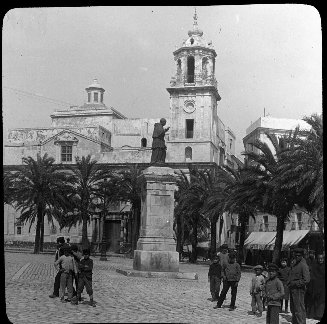 Estatua de un fraile sobre un pedestal en una plaza llena de palmeras. Al fondo, en la esquina de una calle, la torre de una iglesia.