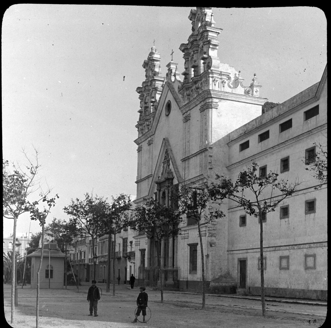 Iglesia barroca con edificios contiguos. Delante tiene un paseo con árboles jóvenes.