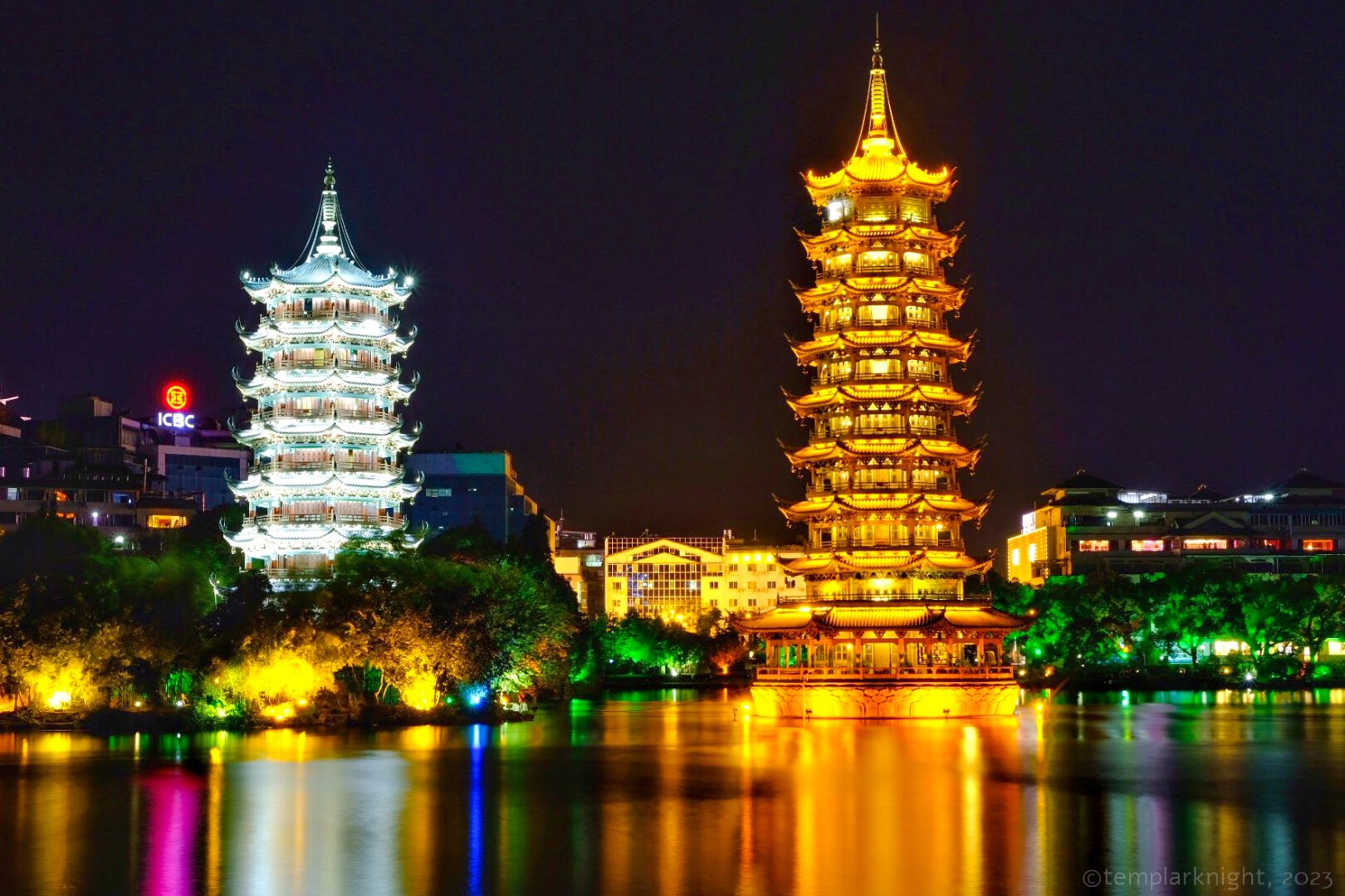 Color photos in landscape format taken at night of two brightly lighted pagoda towers on the side of a small lake in Guilin. Each tower is about 7 -9 stories tall. The one on the right is lighted in yellow-orange and is slightly taller, the one on the left is lighted din light-green. The towers' reflections can be seen on the surface of the lake.