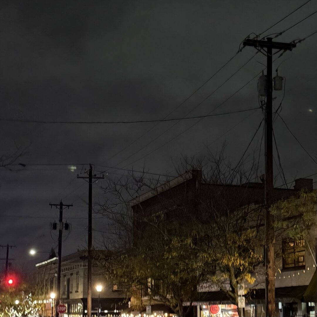 A photo displays a tranquil night scene on a street corner, where buildings, bathed in warm lamplight, create a cozy ambiance. The images low angle captures vibrant green trees, power lines, and the mysterious touch of dark, cloudy skies, evoking a sense of small-town serenity.
