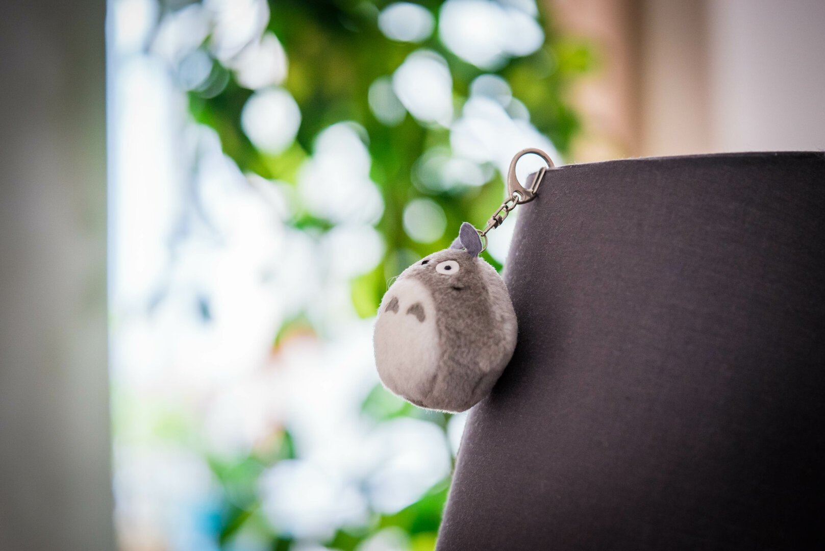 A close-up of a plush Totoro keychain attached to a fabric lampshade. A blurry window with greenery in the background.